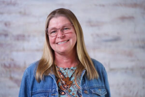 Suzanne Latacz, Realtor® at Ketcham Realty Group in Tallahassee, Flordia, smiling, wearing glasses and a denim jacket, standing in front of a light brick background.